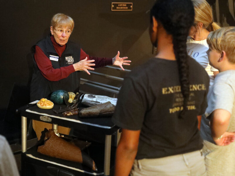 An adult docent interacting with a group of students around a paleo cart of specimens during a museum visit.