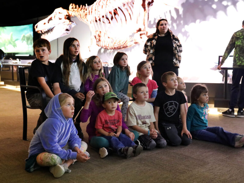 A group of school children sit on the floor during a guided tour at Museum of the Rockies, listening to a presentation in front of a large dinosaur skeleton display.