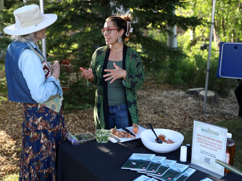 Two women engage in a conversation at the Indigikitchen booth during a Taste of the Rockies event, featuring a table with traditional food samples and educational materials.