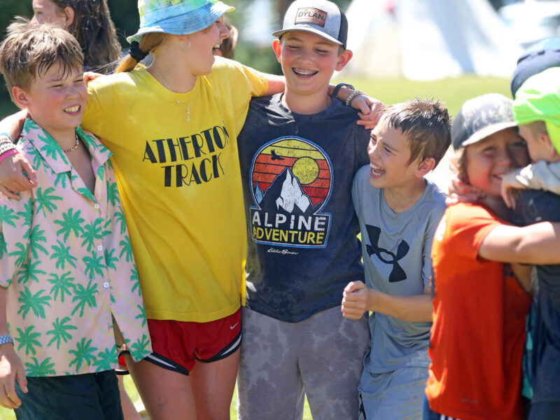 A group of kids smile and laugh together outdoors while wearing colorful t-shirts and hats during a Museum of the Rockies summer camp.