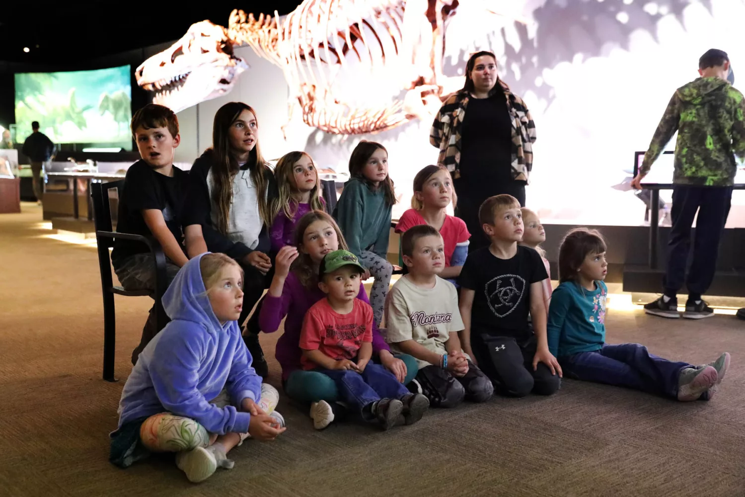 A group of school children sitting on the floor in a museum gallery, looking at a large dinosaur skeleton display during a dinosaur-themed school visit to Museum of the Rockies.