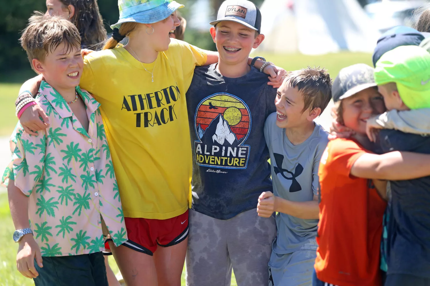 A group of kids smile and laugh together outdoors while wearing colorful t-shirts and hats during a Museum of the Rockies summer camp.