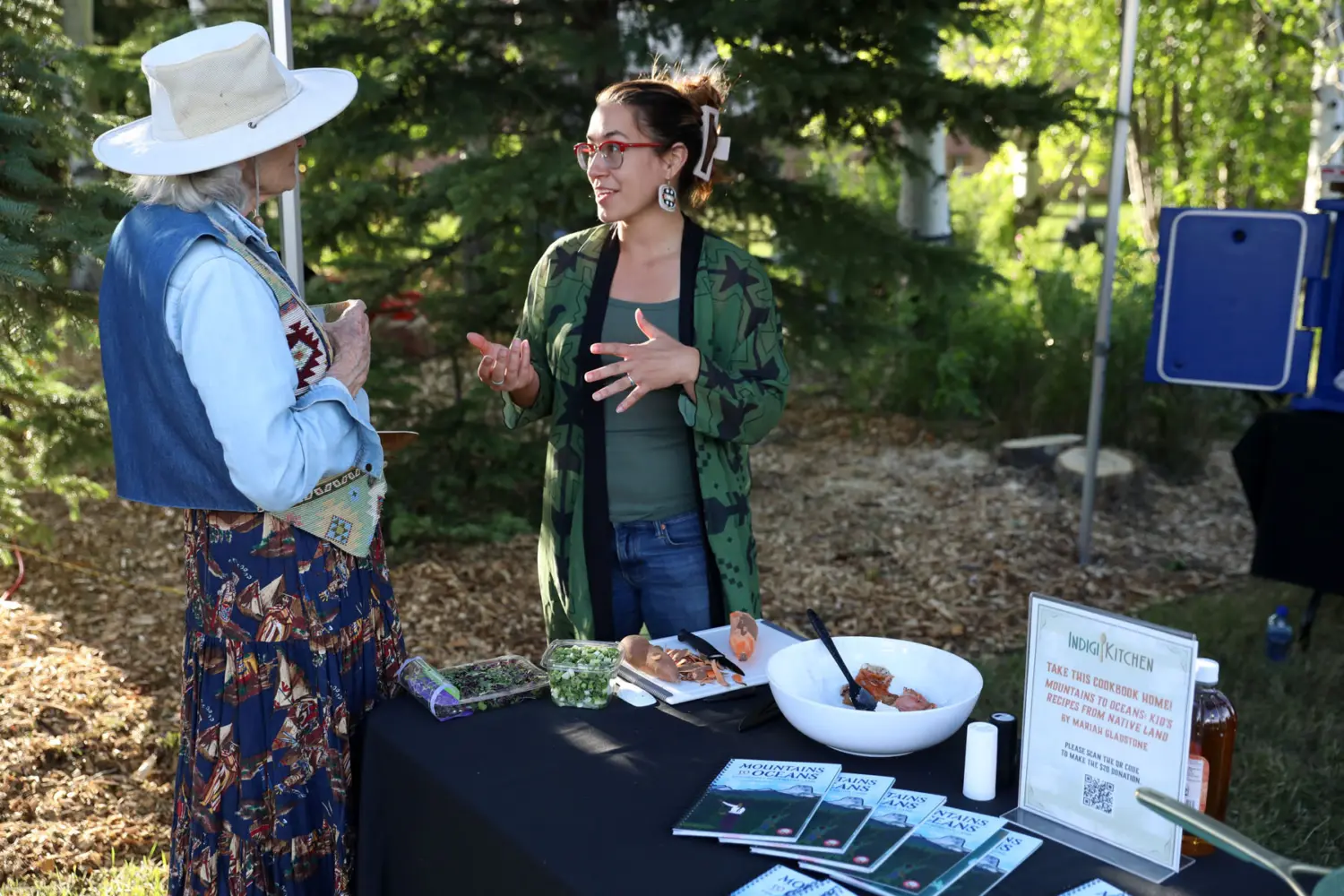 Two women engage in a conversation at the Indigikitchen booth during a Taste of the Rockies event, featuring a table with traditional food samples and educational materials.