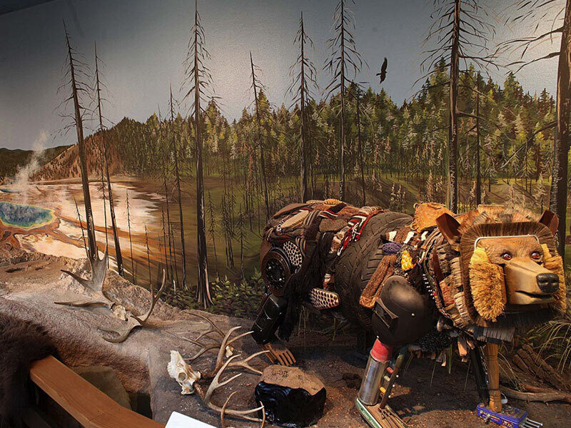 A sculpture of a grizzly bear and cub handcrafted from repurposed household materials, displayed in an immersive forest and geothermal environment within the "Explore Yellowstone" exhibition.