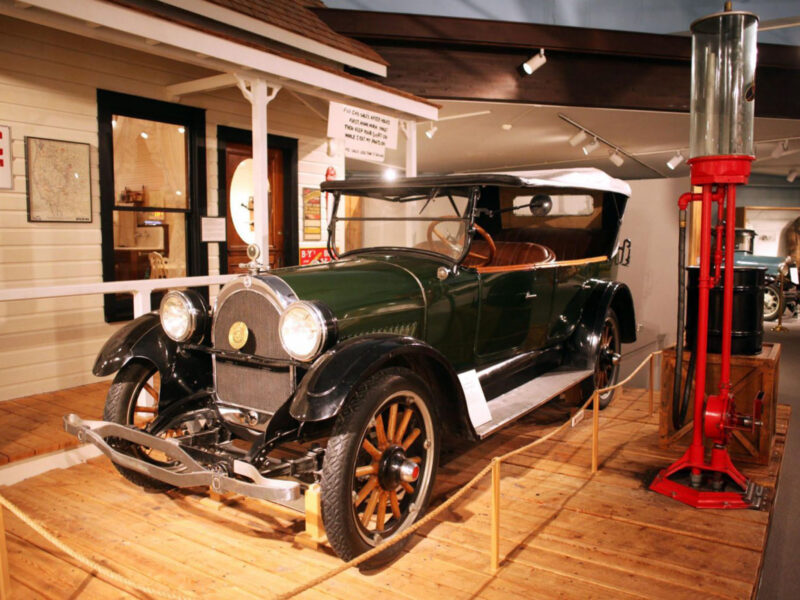 A vintage 1922 Oldsmobile Touring Car with a green body, black fenders, and wooden-spoke wheels, displayed on a wooden boardwalk in front of a replica gas station within the Paugh History Hall exhibit.