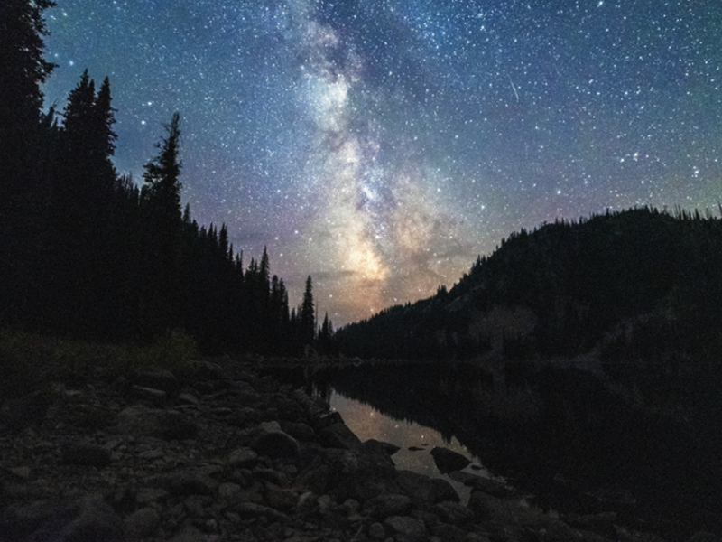 A stunning nighttime photograph of the Milky Way arching over a dark mountain landscape and a still body of water.