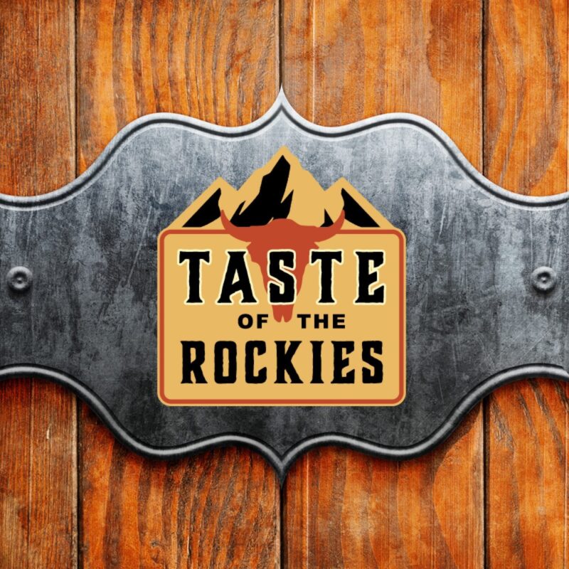 A logo for "Taste of the Rockies" featuring a mountain graphic on a decorative metal badge, mounted against a vertical wooden plank background.