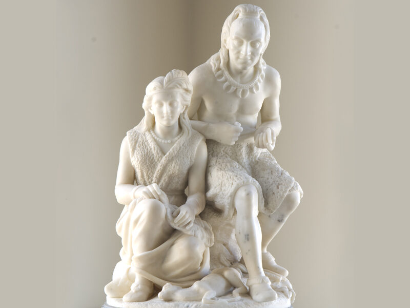 White marble sculpture titled "The Wooing of Hiawatha" by Edmonia Lewis, depicting two seated figures in detailed traditional attire on a circular base.